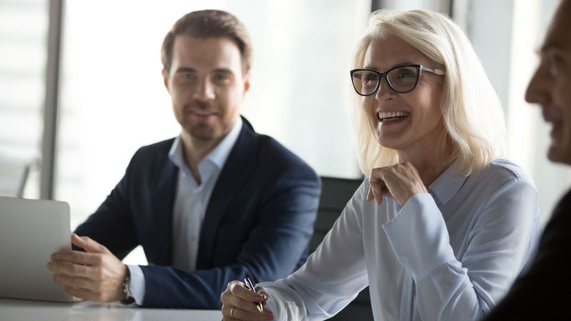 Das Bild zeigt eine Besprechung, bei der 3 Personen zu sehen sind. Eine Frau ist im Fokus gezeigt ist und an ihrer Seite sitzen zwei Herren.